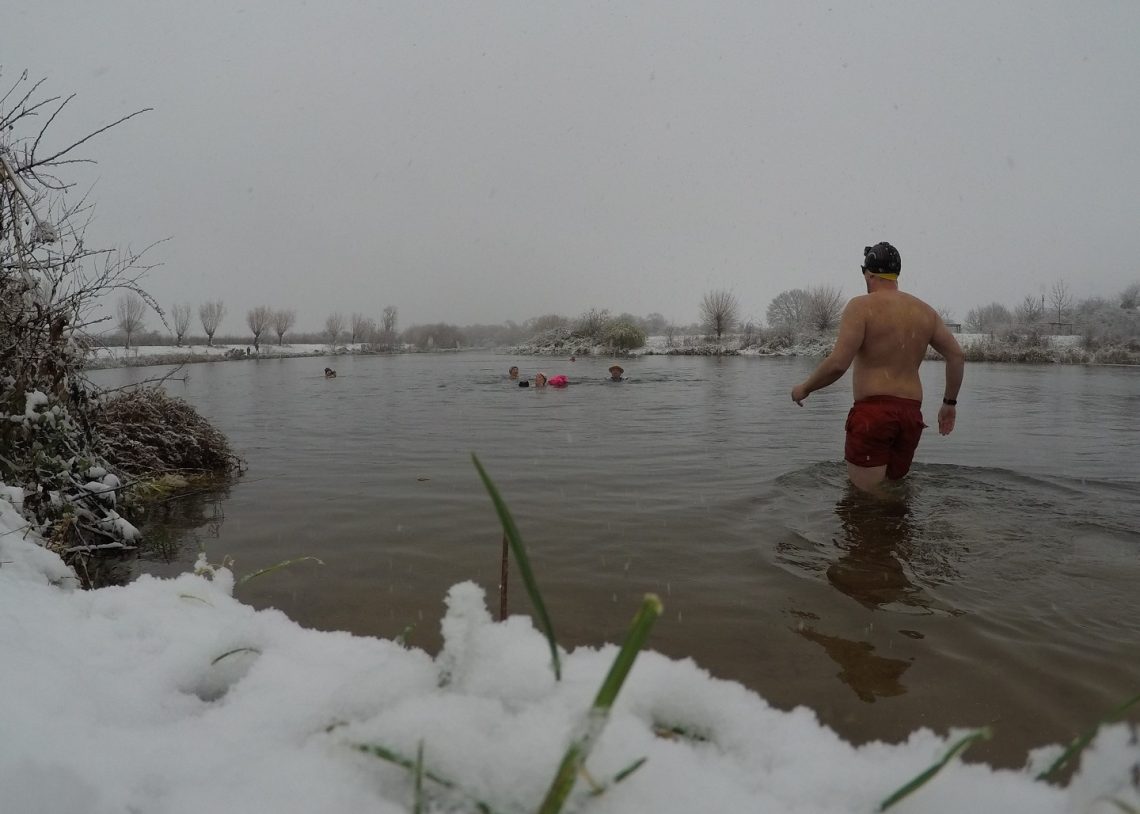 A man standing knee deep in water facing away from the camera. Other swimmers in the distance. Snow on the ground in the foreground.