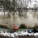 Two people standing waist deep in a river, facing away from the camera. Snow on the ground.
