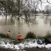 Two people standing waist deep in a river, facing away from the camera. Snow on the ground.